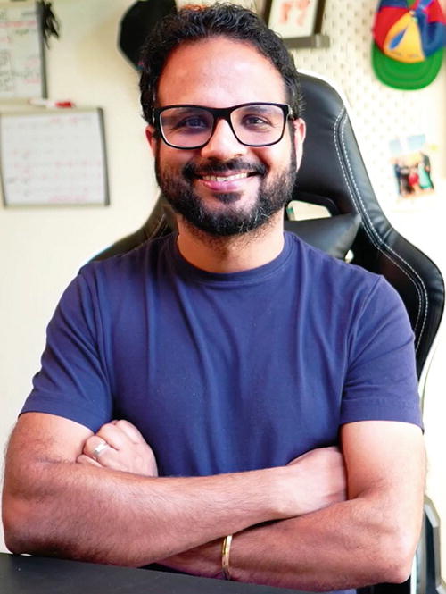 A person with glasses and a beard is smiling while sitting in a chair with arms crossed. They are wearing a blue shirt. In the background, there is a whiteboard with writing, a colorful cap, and a few framed pictures on the wall. The setting appears to be an office or study space.