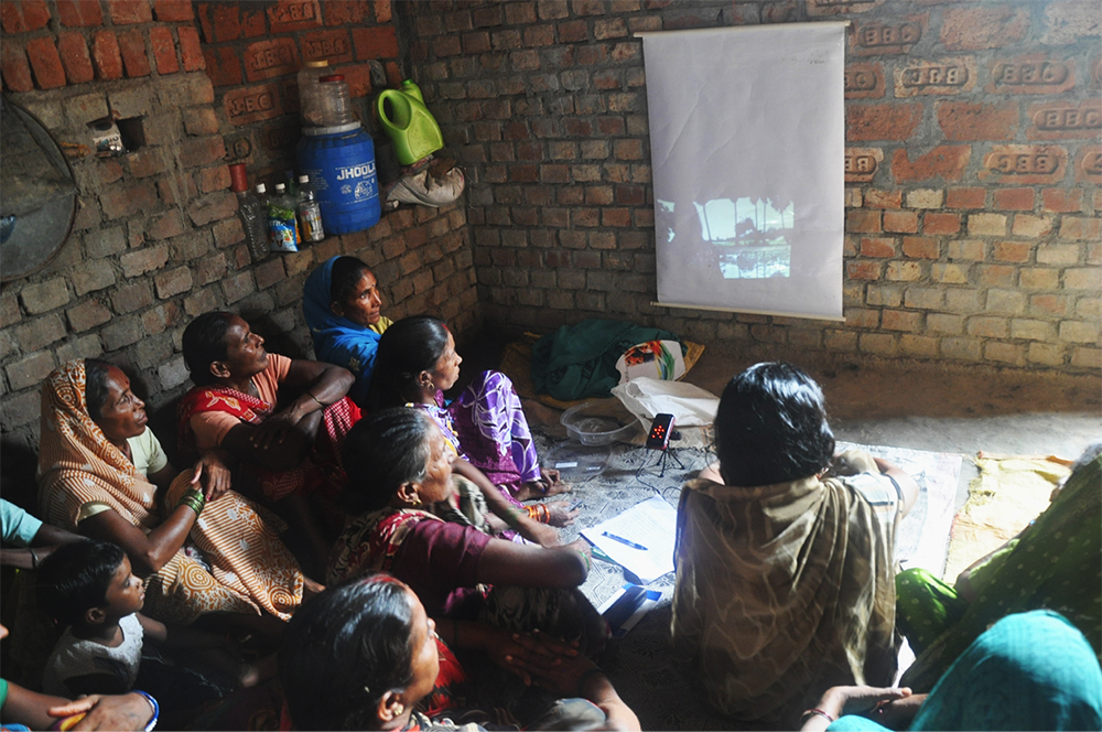 Small brick room full of people watching a video being projected on the wall.
