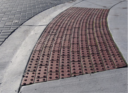 An arc of puce-colored ADA warning pads between a light grey concrete sidewalk and a dark grey brick-patterned surface.
