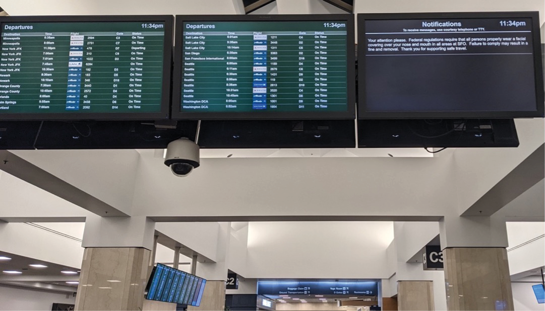 An airport concourse, with three large ceiling-mounted monitors displaying plane departures and other notifications.
