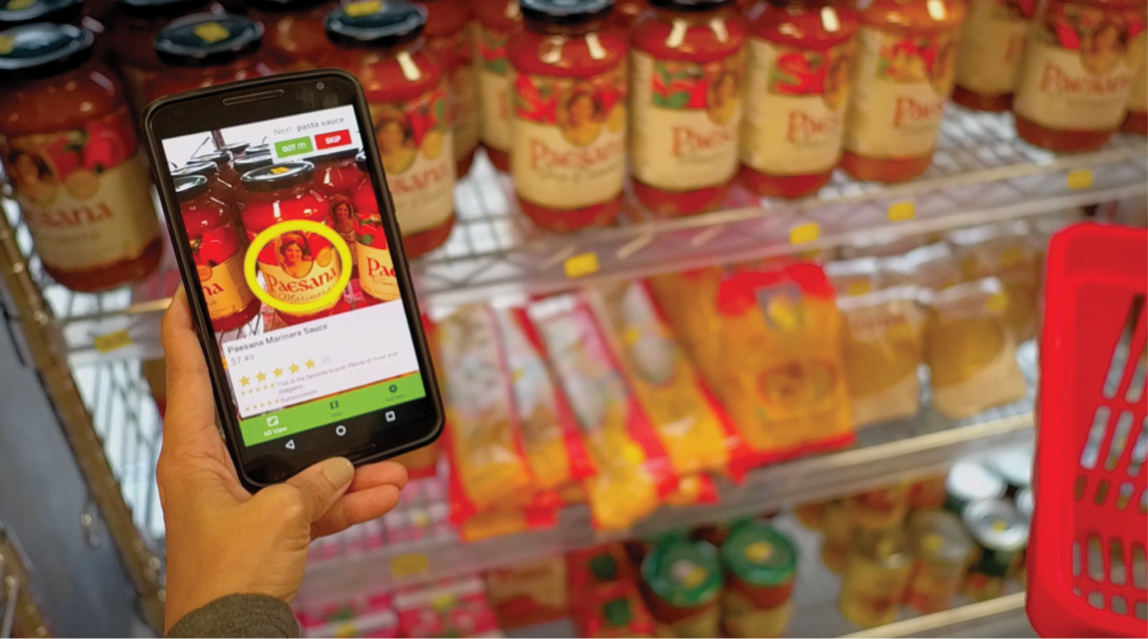Hand holding a smartphone showing a live photo of sauce jars in a grocery aisle. On the device display, a yellow circle centers on the photo.