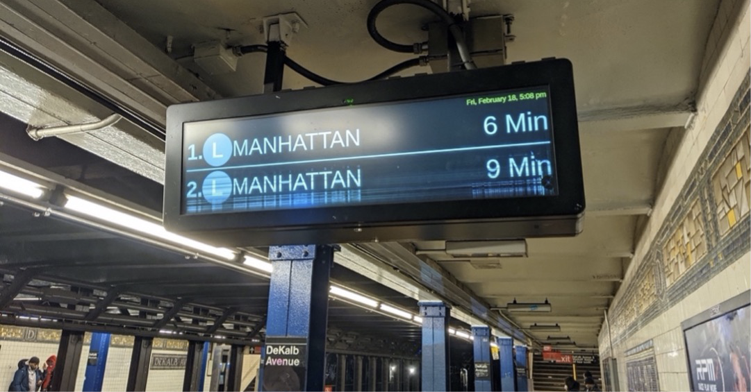 A ceiling-mounted digital sign in a subway station shows two wait times for the next train.