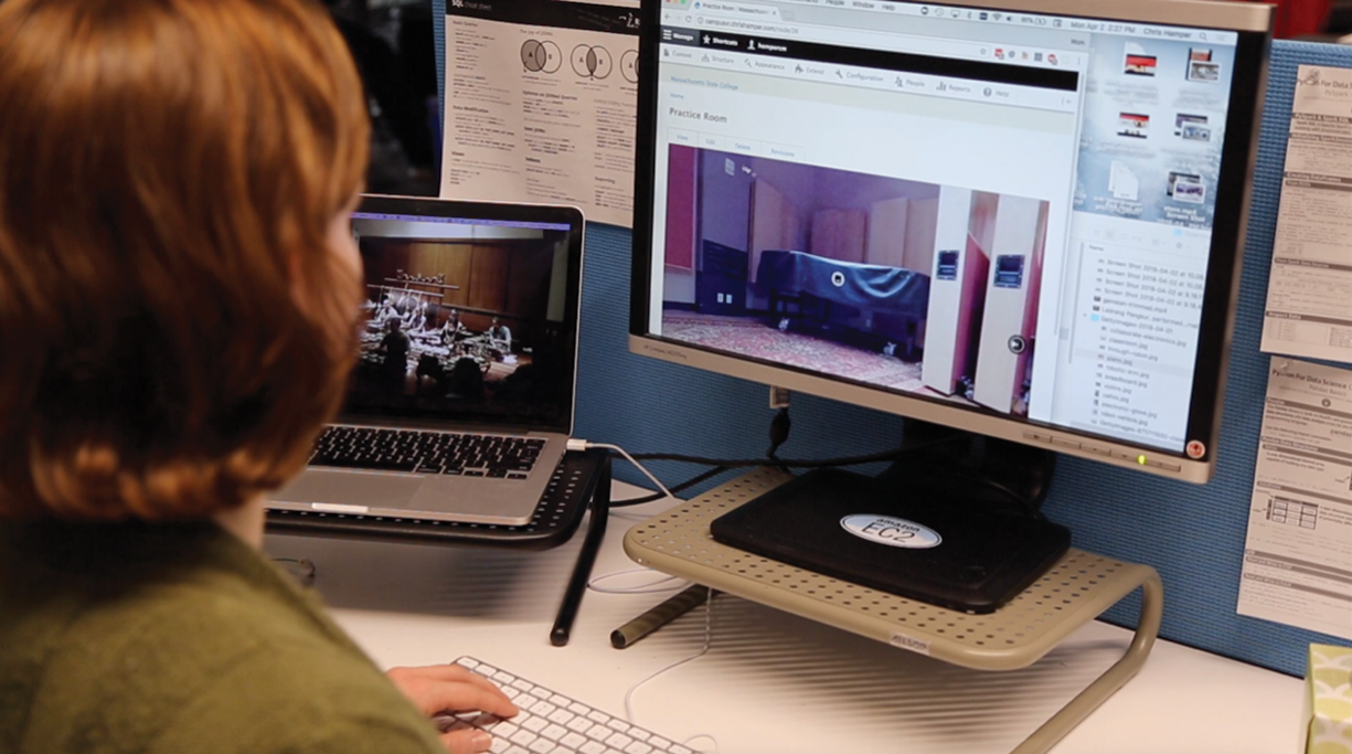 A person with red hair uses a white keyboard while sitting at a desk with a laptop connected to a monitor.