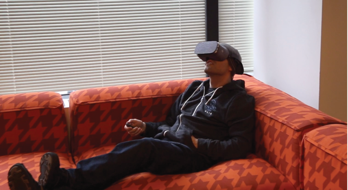 A man wearing a VR headset reclines on an orange houndstooth-patterned couch.